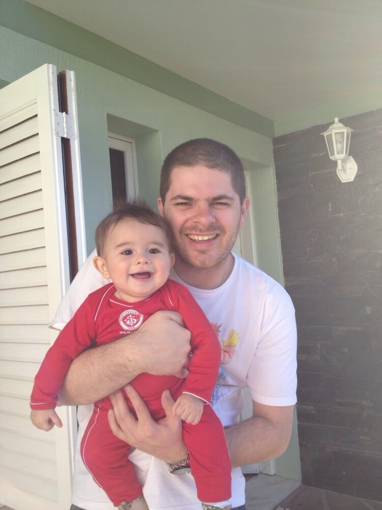Tio Baboo segurando o Deds bebê, de macacão vermelho do Internacional, na varanda de casa