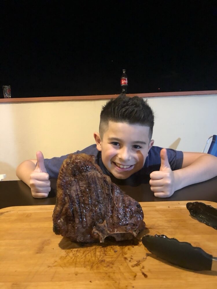 Deds com os dois polegares para cima atrás de um T-bone grelhado