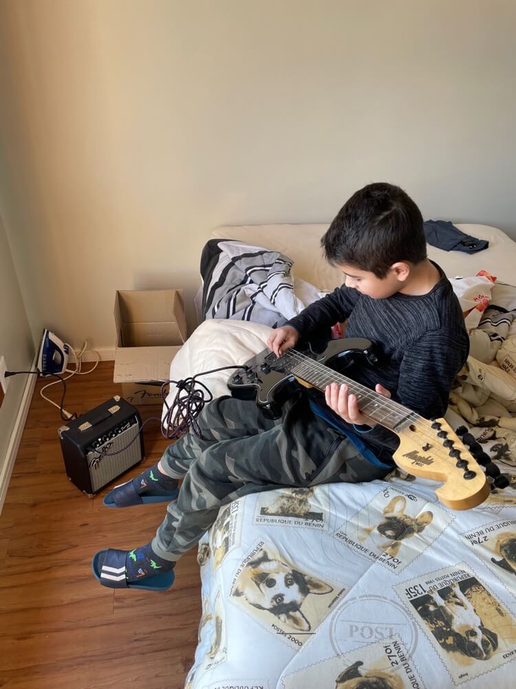 Deds sentado no quarto de visitantes do antigo apartamento do Tio Baboo, concentrado tocando pela primeira vez a guitarra dele, com o amplificador ligado ao lado