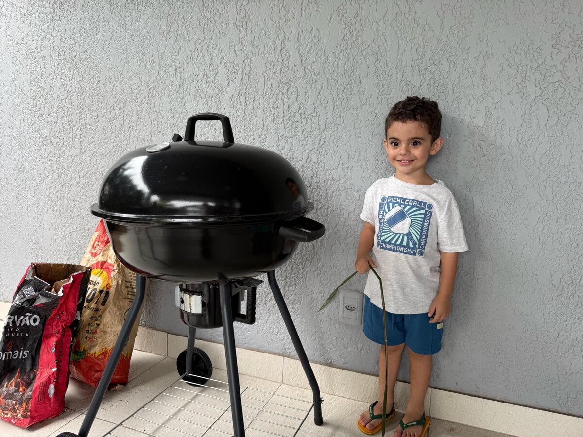 Chico sorrindo encostado na parede ao lado de uma churrasqueira bola preta e dois sacos de carvão
