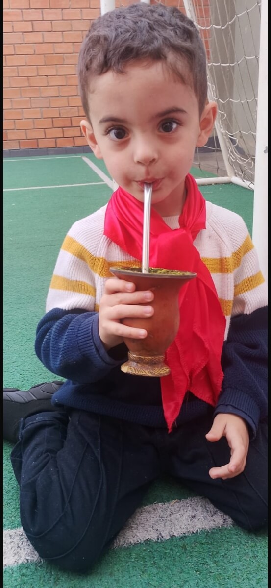 Chico de lenço vermelho no pescoço, sentado na grama sintética, tomando chimarrão na cuia