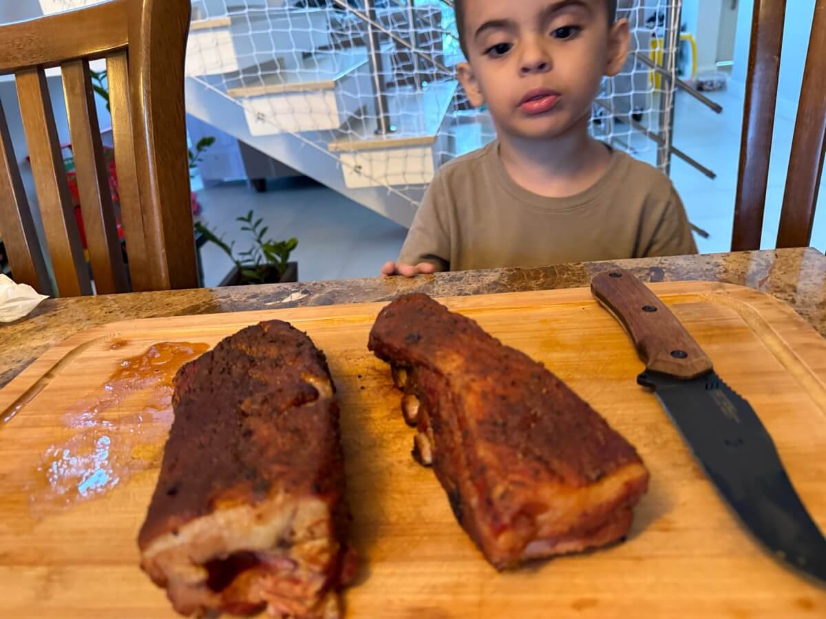 Chico olhando atento para duas peças de carne defumada sobre a tábua, com a faca ao lado