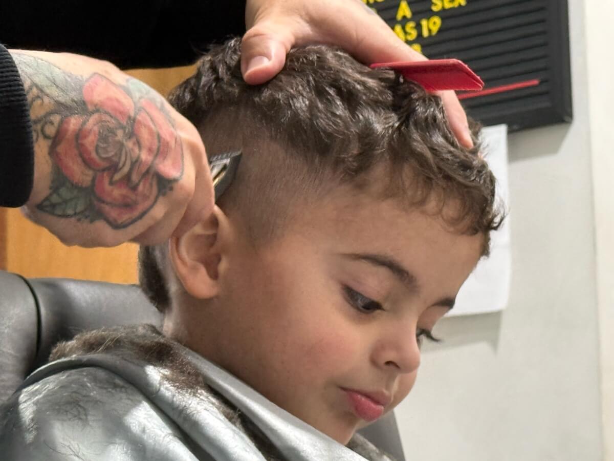Chico cortando o cabelo, de capa, enquanto o barbeiro acerta a lateral com a máquina