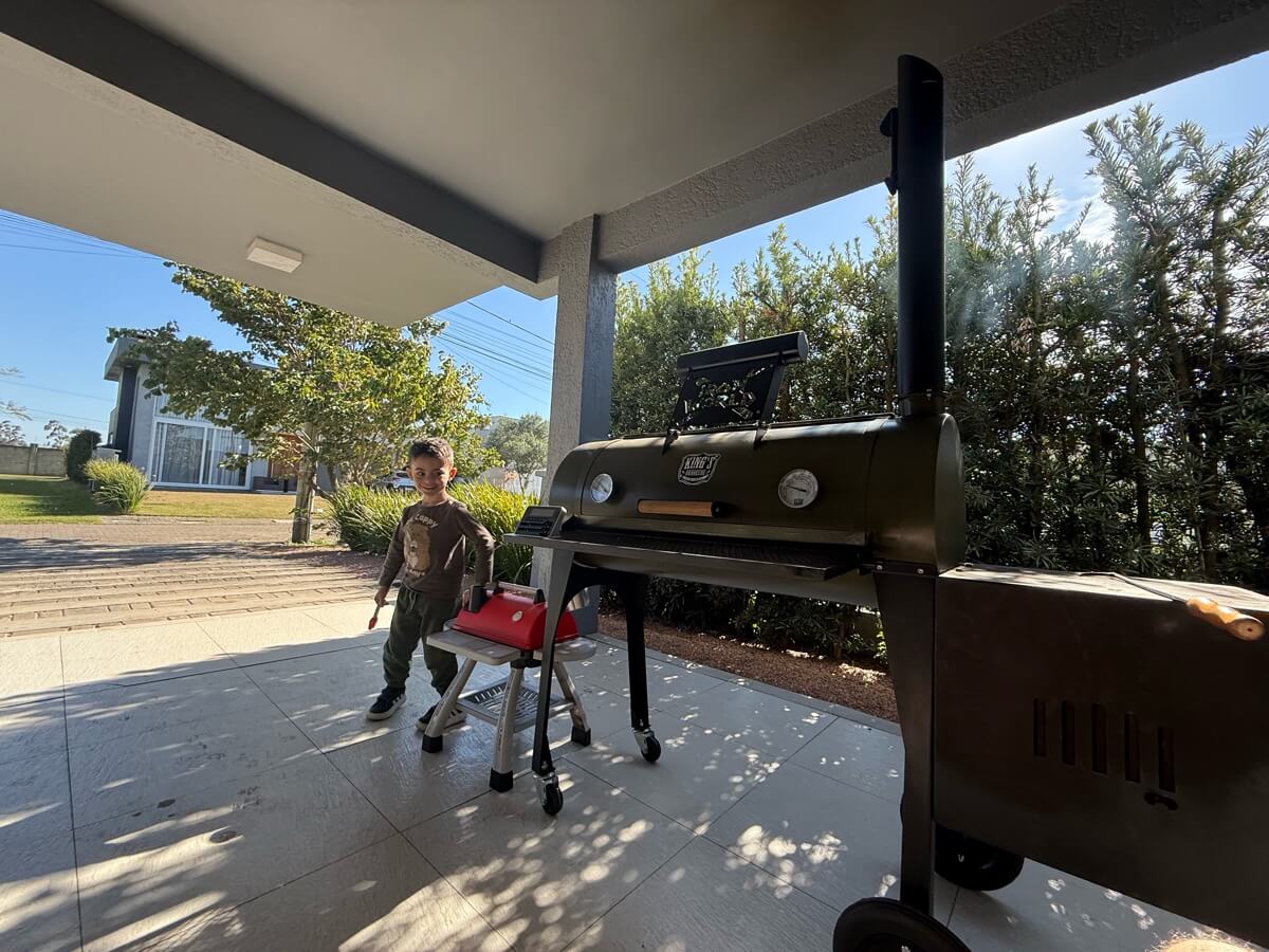 Chico na varanda com a churrasqueira de brinquedo vermelha, ao lado do defumador de verdade soltando fumaça